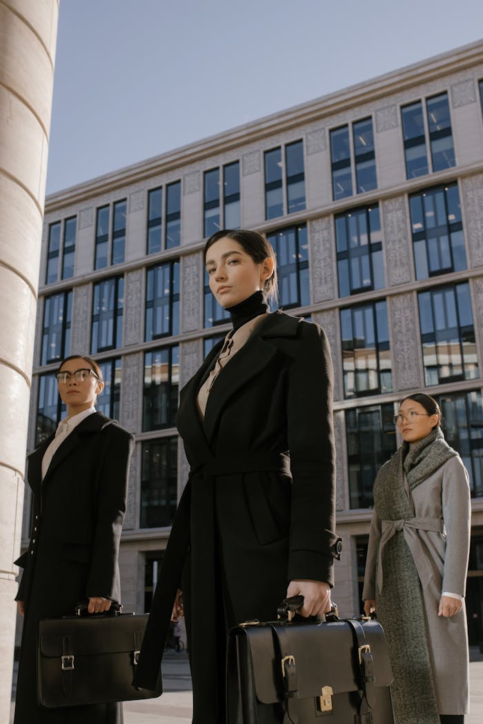 Maison Three professional women in coats carrying briefcases stand confidently in front of a modern office building.